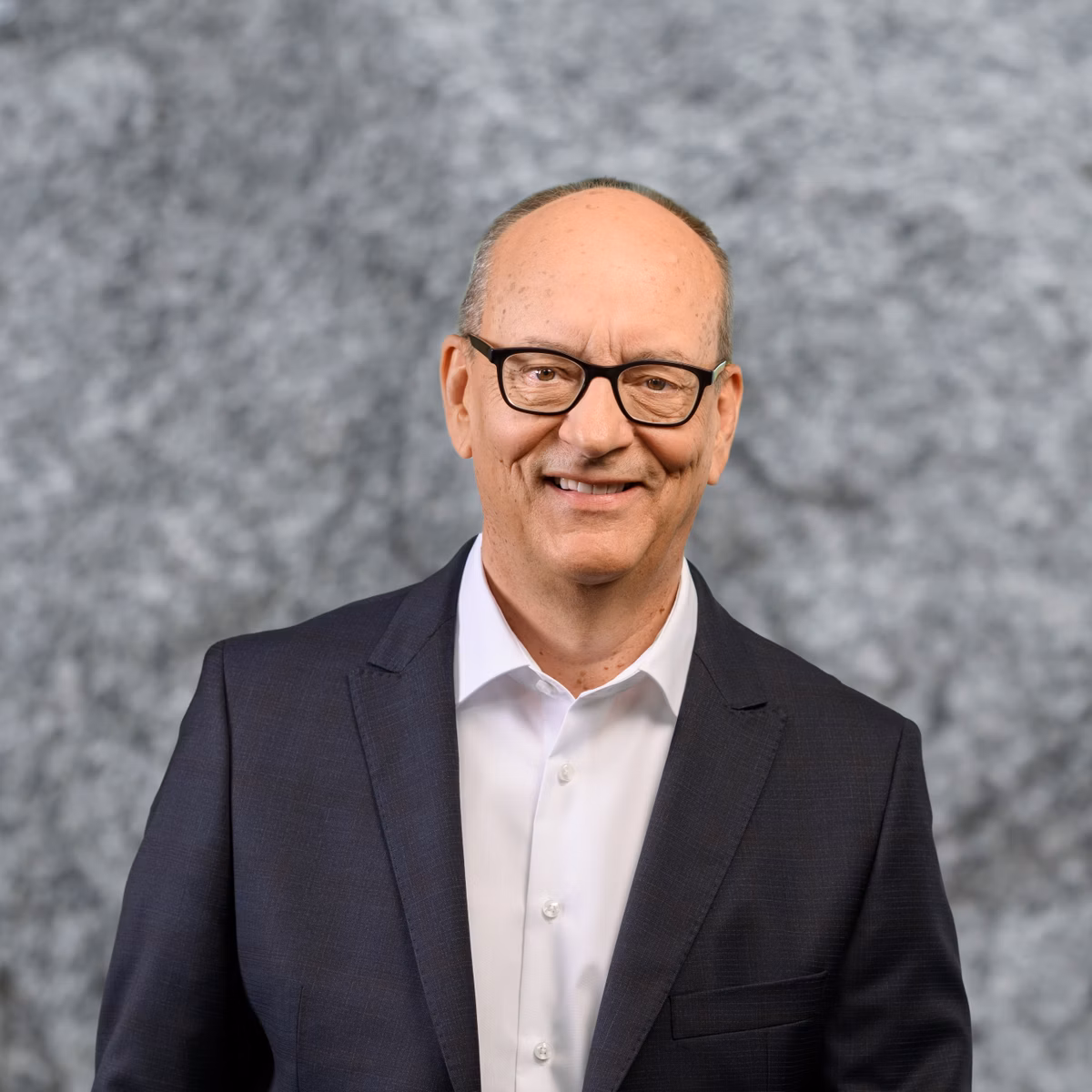 Portrait of a smiling man with glasses, a white shirt and a dark suit against a grey background