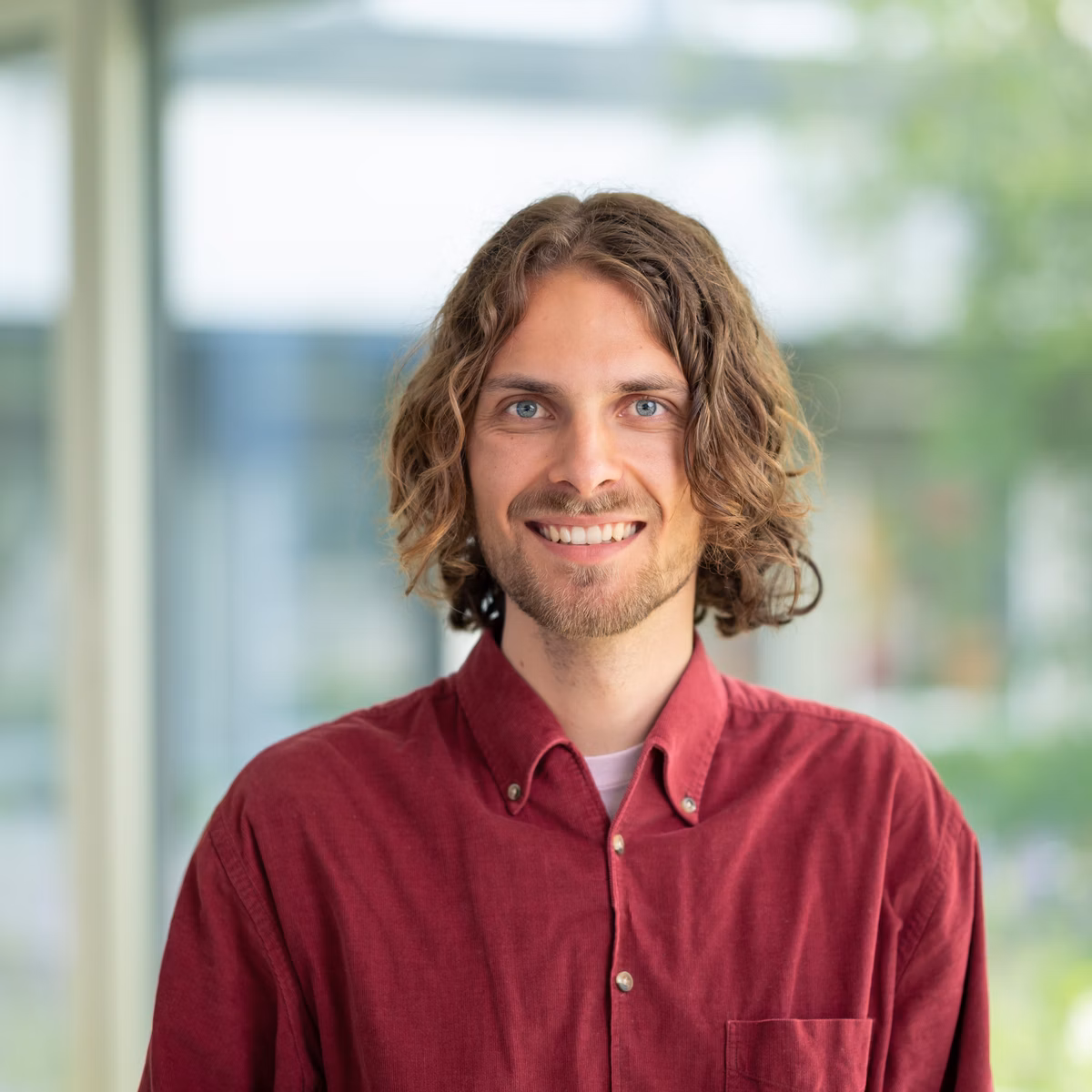 Porträt eines lächelnden Mannes mit schulterlangen Haaren und rotem Hemd vor grossen Fenstern