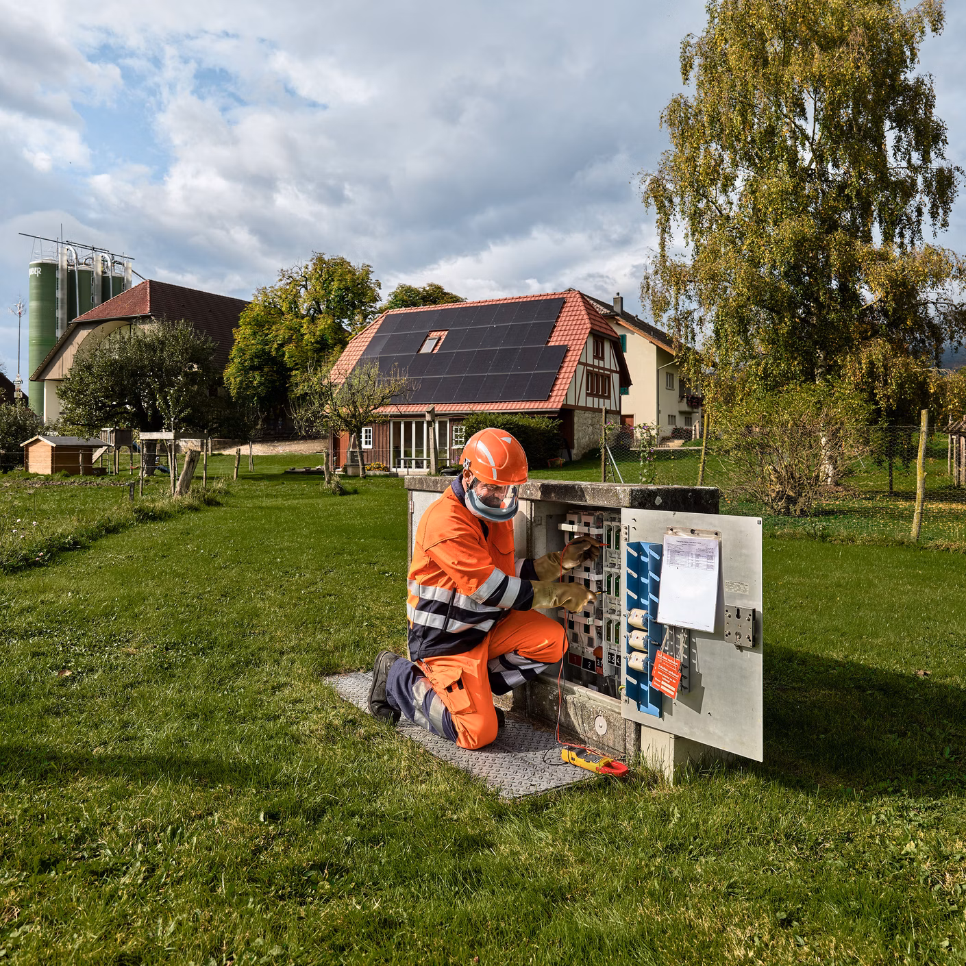 Person in orangefarbener Arbeitskleidung arbeitet an einem offenen Verteilerkasten auf einer Wiese in einem Wohngebiet.
