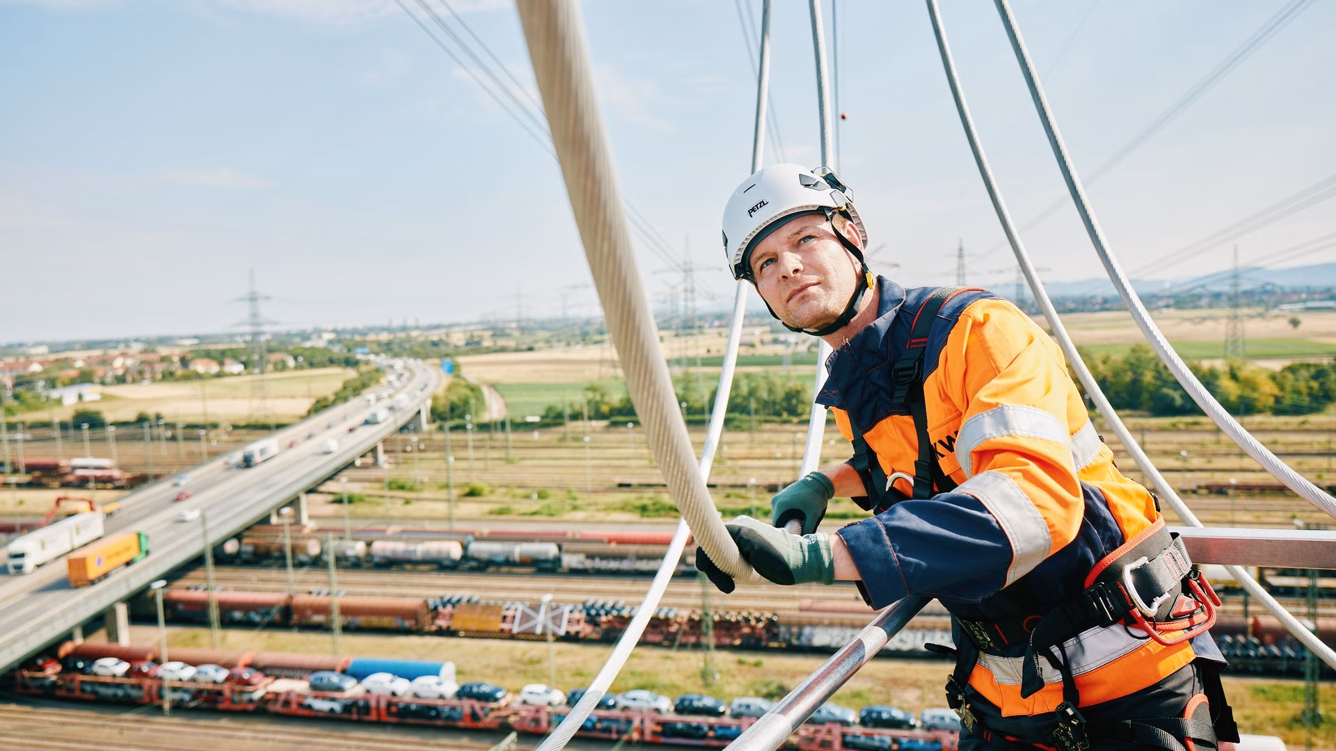 Mitarbeitender in Schutzkleidung und Kletterausrüstung auf einem Hochspannungsmast; im Hintergrund sind Gleisanlagen, Verkehrswege und weitere Strommasten zu sehen.