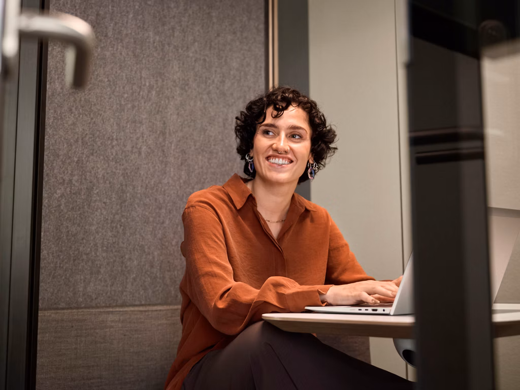 Frau sitzt lächelnd an einem Tisch und arbeitet an einem Laptop in einem modernen Innenraum.
