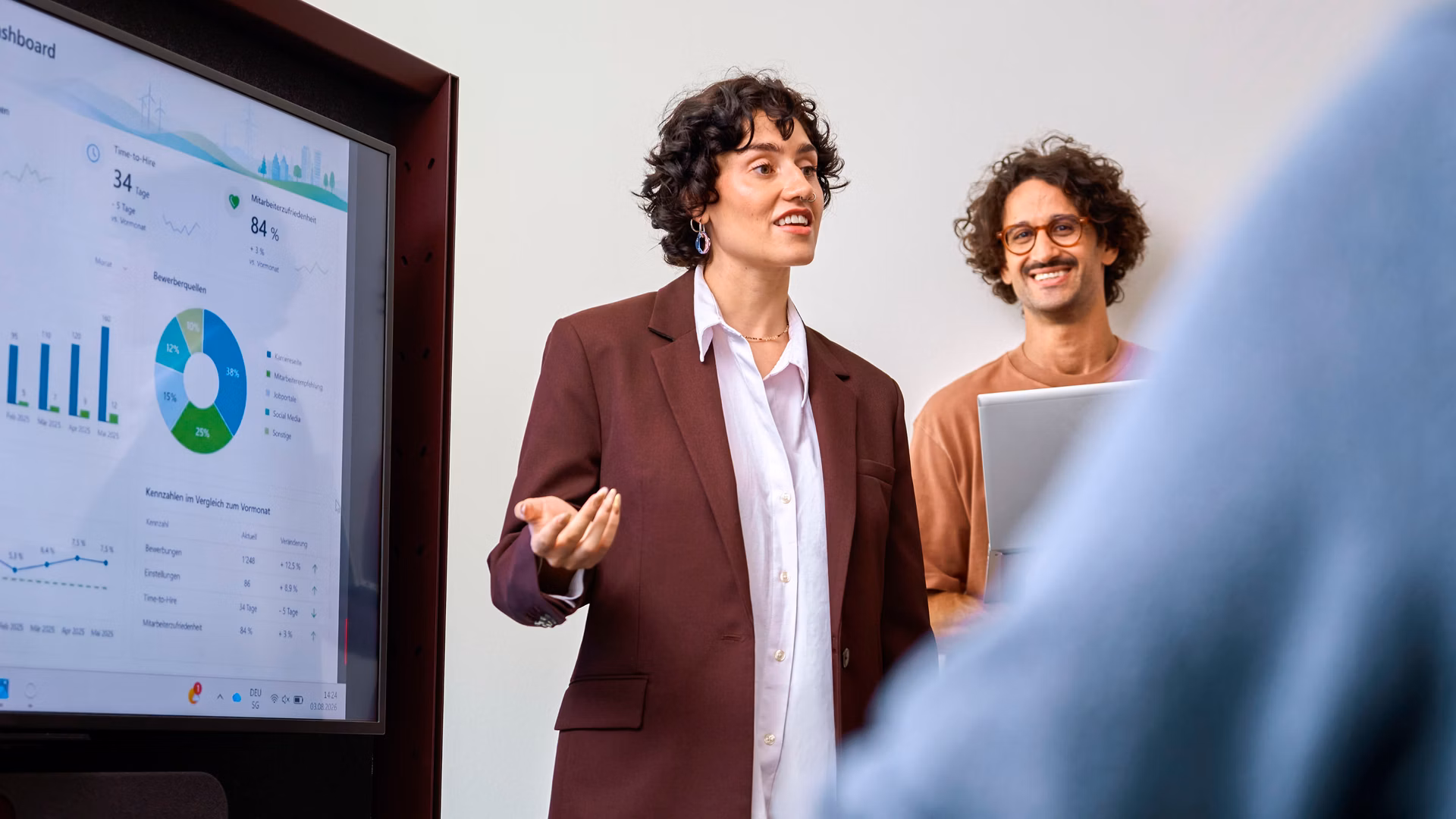 Two people present key figures on a large screen in a meeting room.