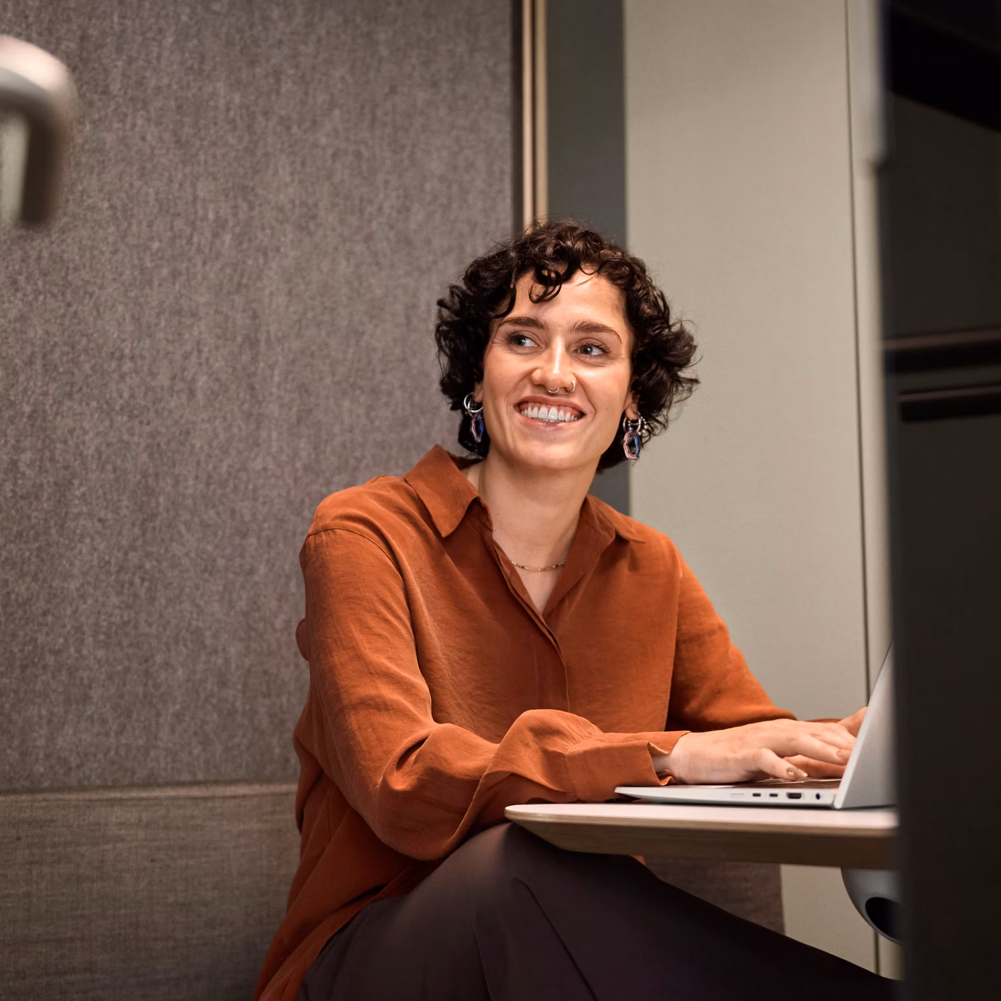 A woman sits smiling at a table and works on a laptop in a modern interior.