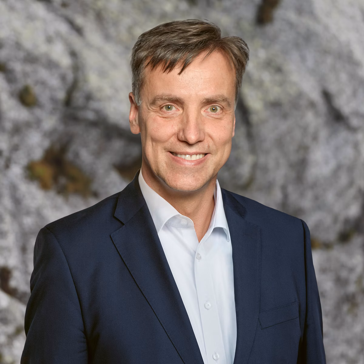 Man in a dark suit in front of a grey rock face