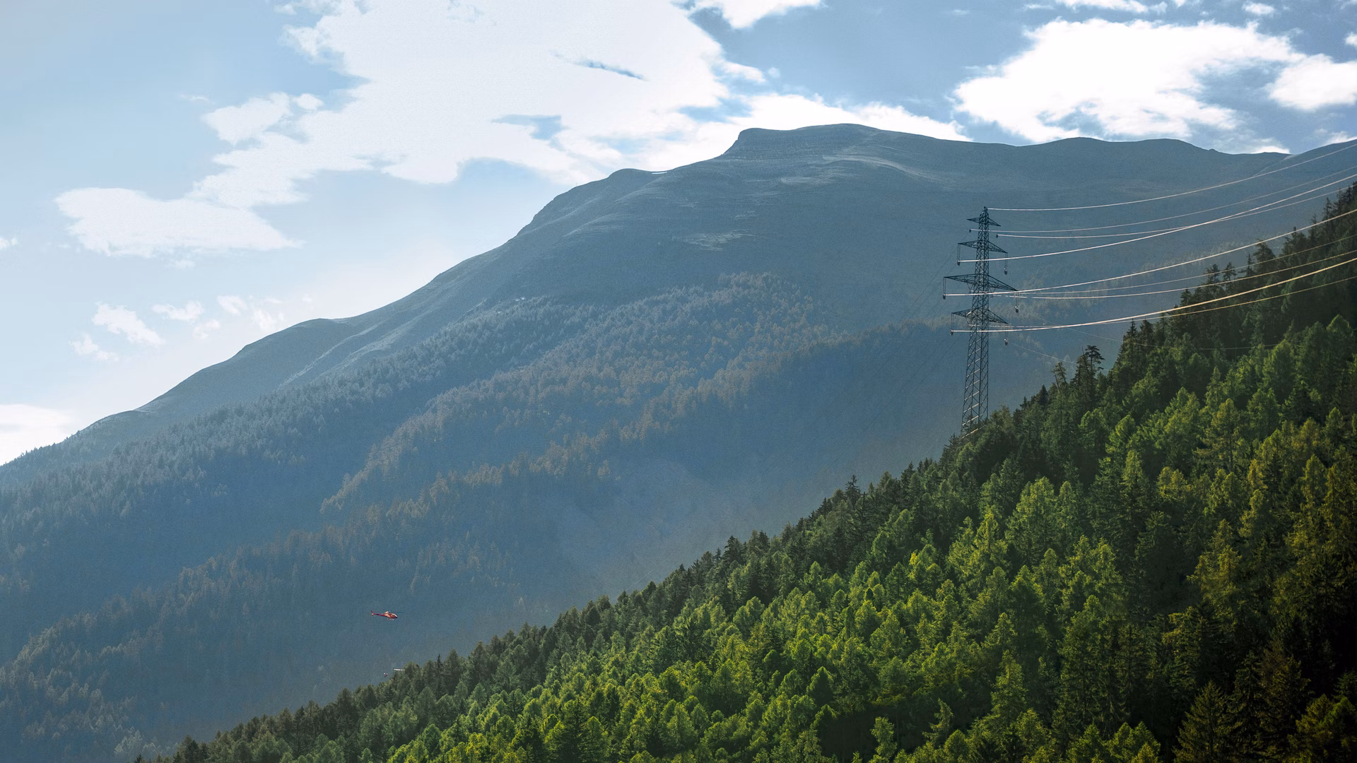 Strommast mit Leitungen an einem bewaldeten Berghang vor Bergkulisse.