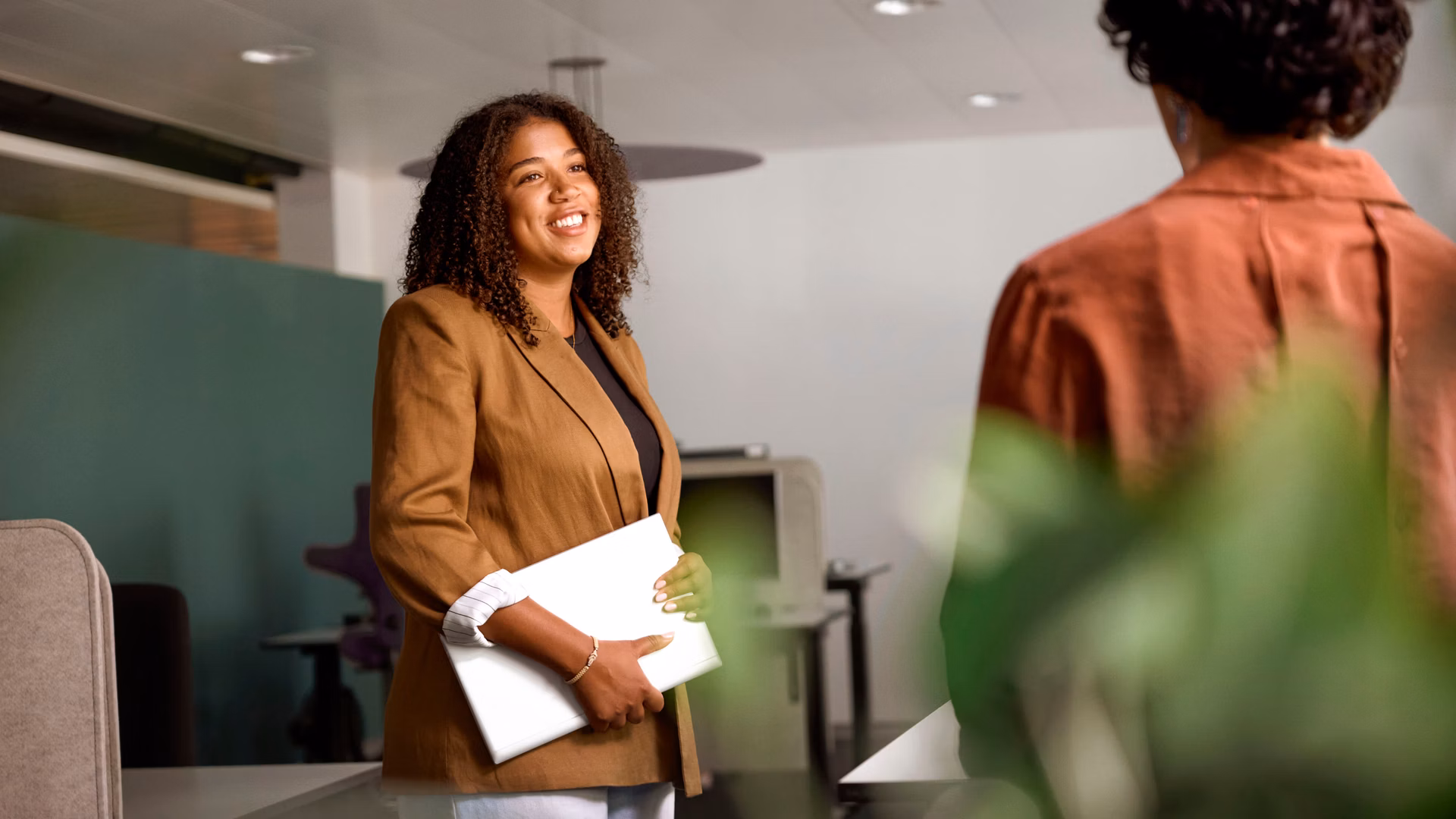 Two people talking in a modern office, one woman holding a notebook in her hand.