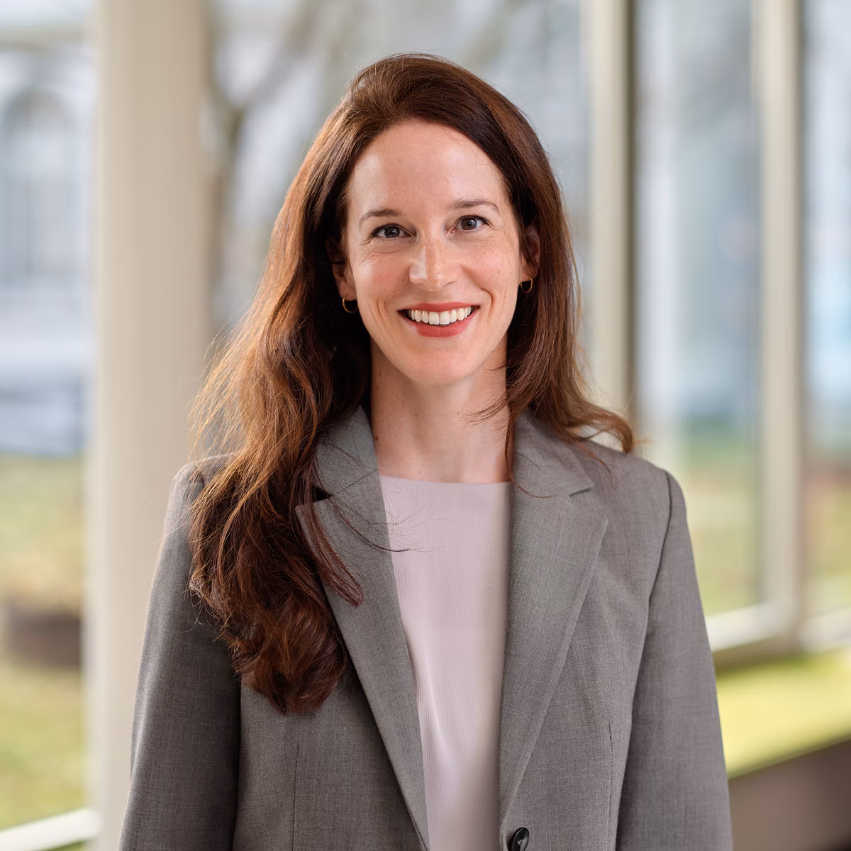 Portrait of a woman with long red hair in a grey blazer in front of large windows