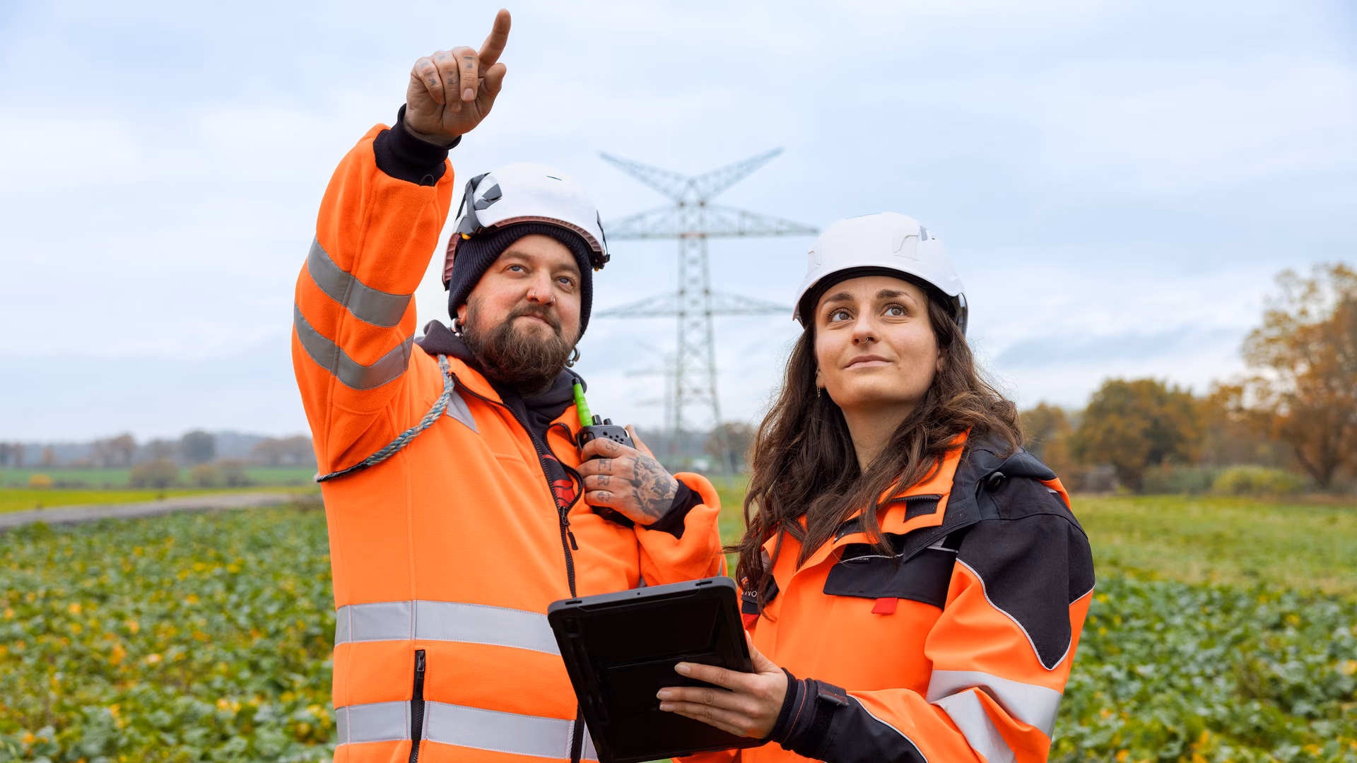 Zwei Personen in orangefarbener Schutzkleidung und Helmen stehen mit einem Tablet auf einem Feld vor einem Strommast.