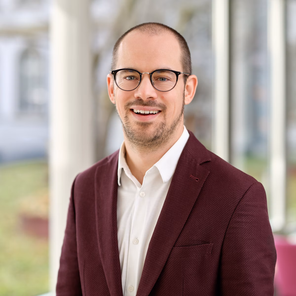Man wearing glasses, a burgundy jacket and a white shirt in a bright interior