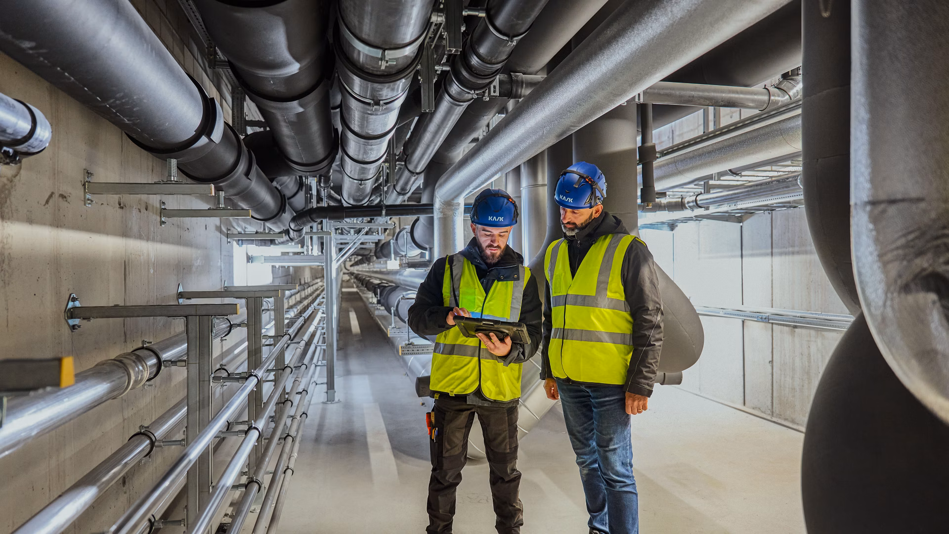 Zwei Arbeiter mit Schutzhelmen und Warnwesten stehen in einem Technikraum mit Rohrleitungen.