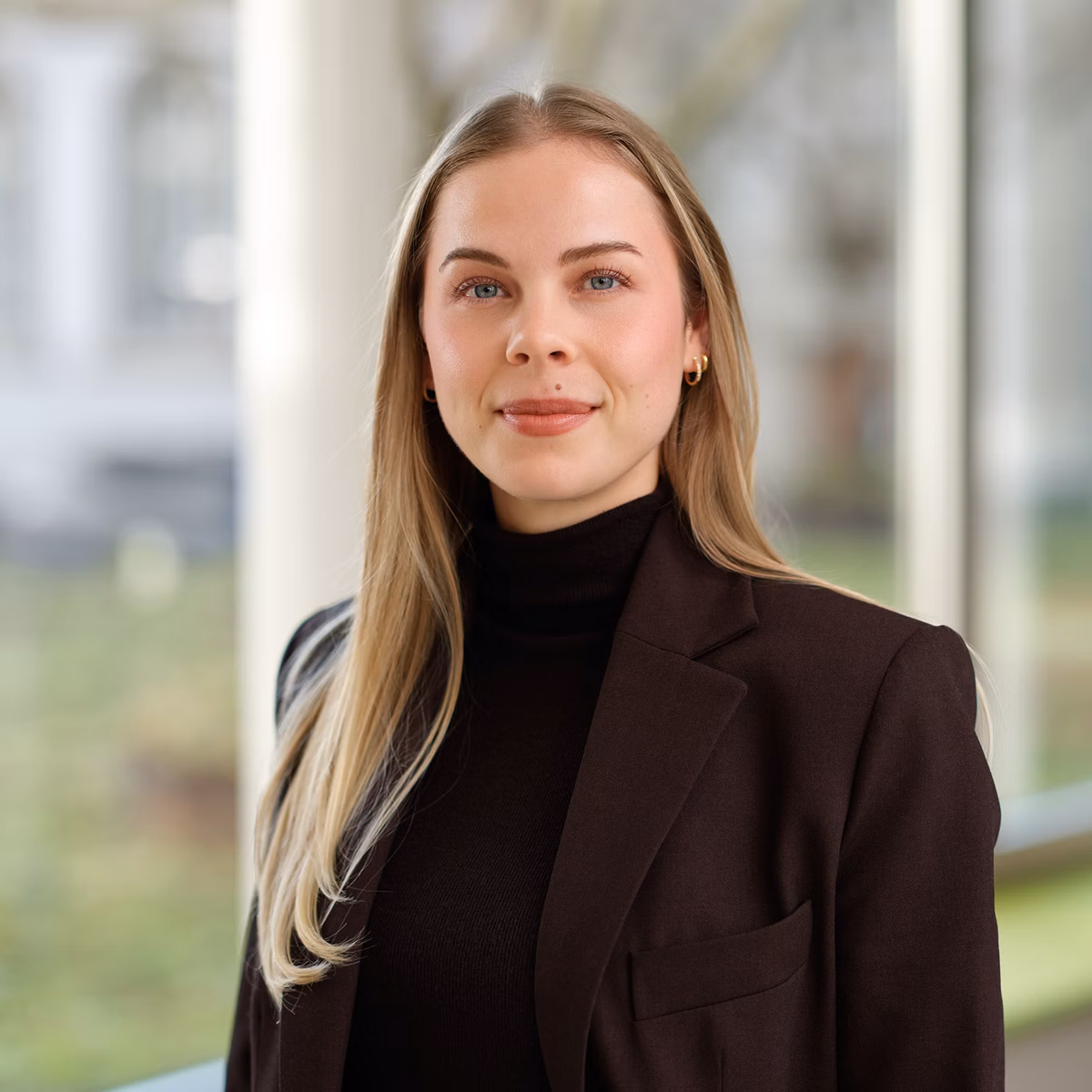 Young woman in a dark blazer in front of large windows.