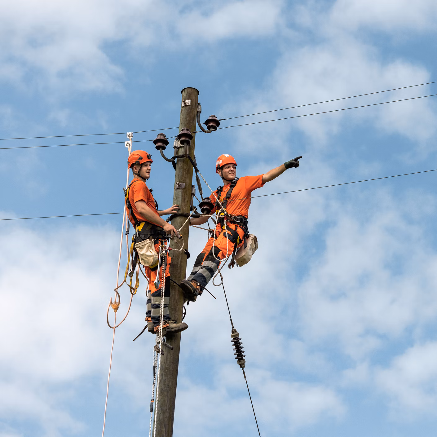 Two technicians with climbing equipment are working on an electricity pylon beneath overhead power lines.