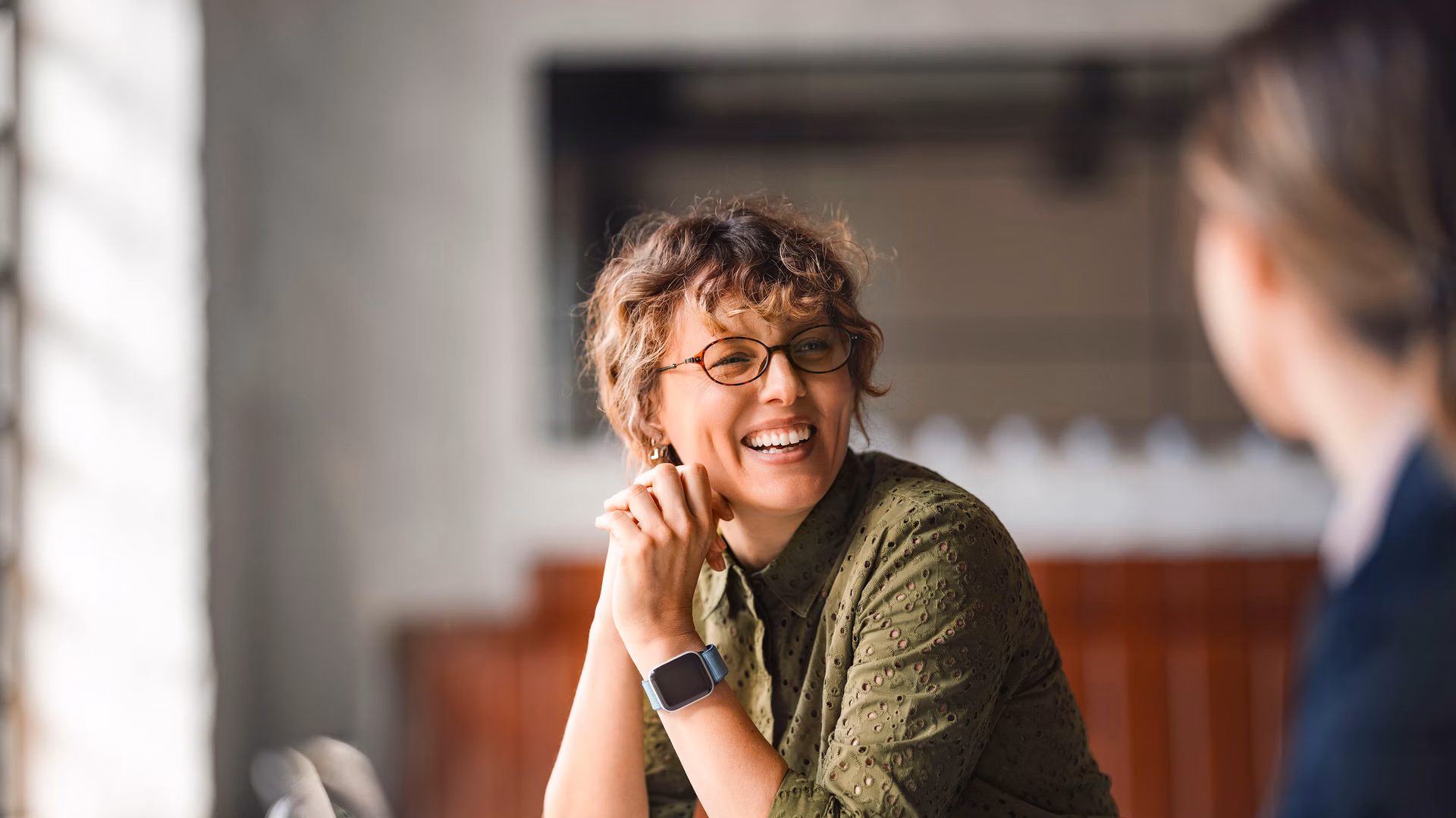 Two people in conversation at a table, a woman wearing glasses smiles.
