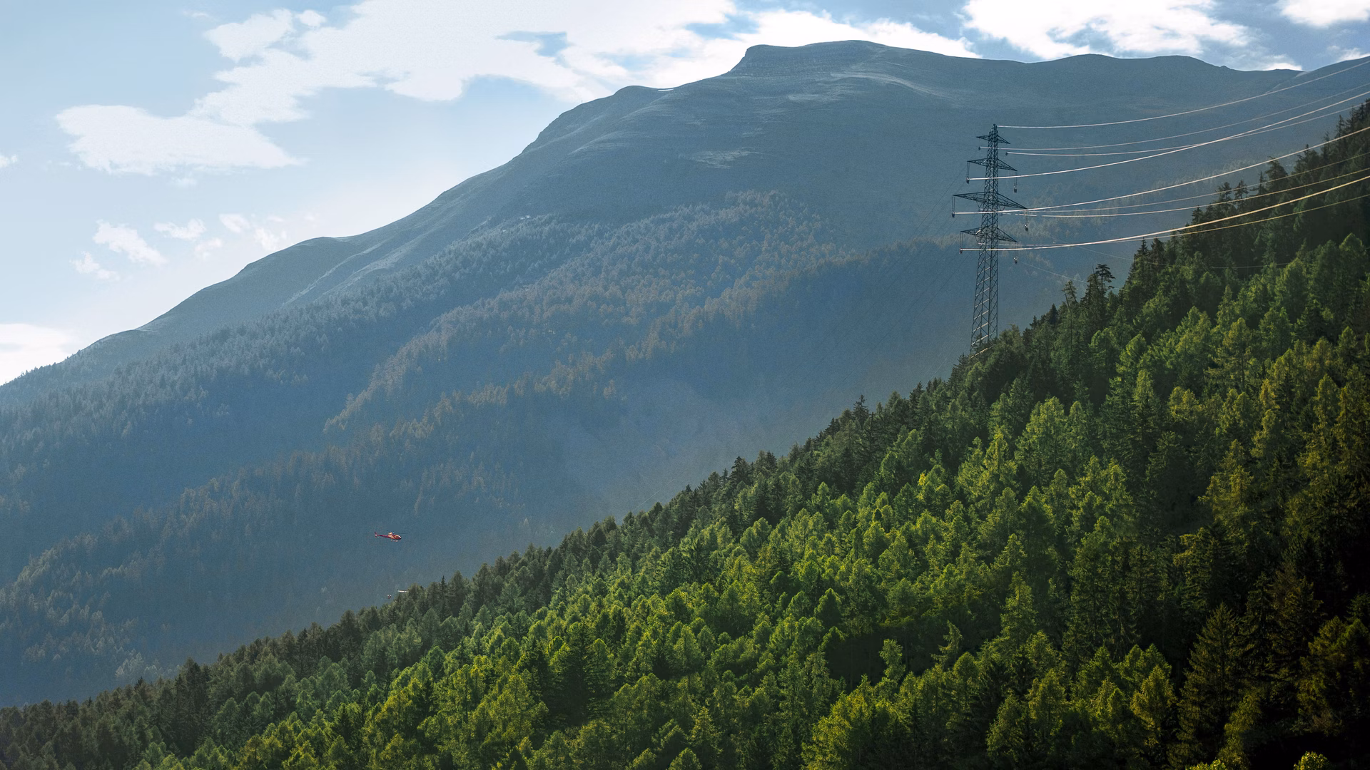 Strommast mit Leitungen an einem bewaldeten Berghang vor Bergkulisse.
