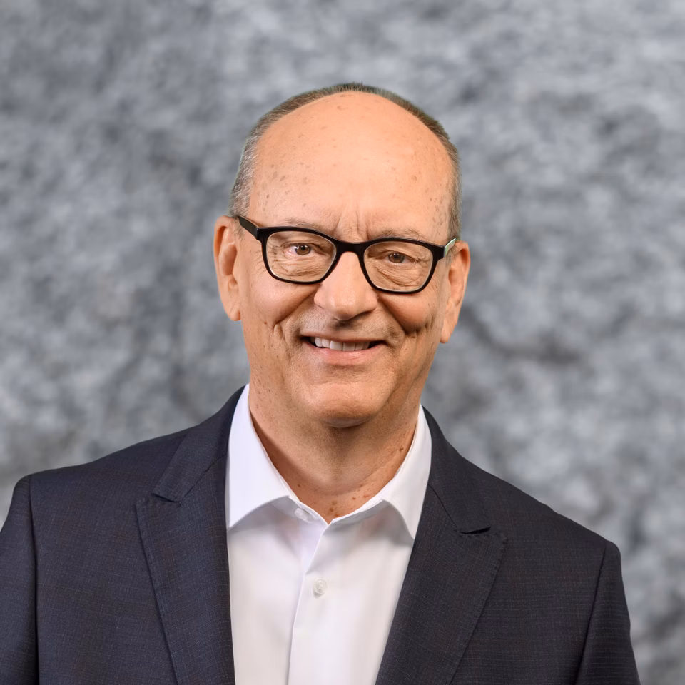 Porträt eines lächelnden Mannes mit Brille, weissem Hemd und dunklem Anzug vor grauem Hintergrund
